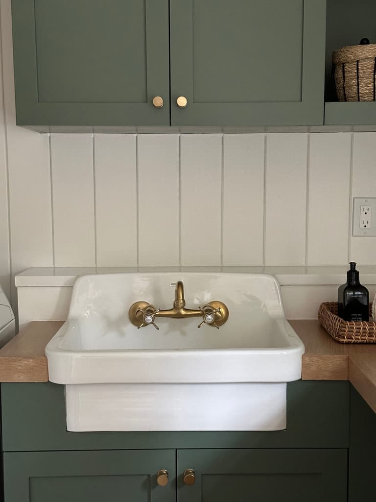 Green shaker laundry cabinetry and vertical shiplap around an apron-front sink with a brass bridge tap in a Mendham, NJ renovation by Michael Robert Construction